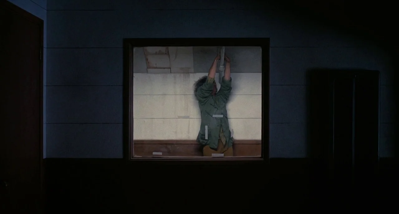 Inherent Vice (2014) movie still: medium — A dark, dim room frames a person through a rectangular observation window. The person stands inside the…
