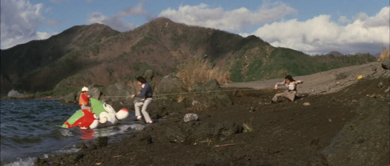 Godzilla Vs Megalon (1973) movie still: wide — A rocky volcanic lakeshore sits beneath a range of mountains under a cloudy sky. Three individuals…