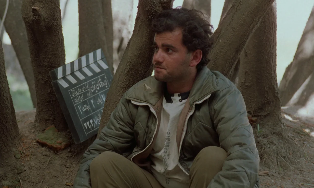 Through The Olive Trees (1994) movie still: medium — A man sits on the ground resting his back against a thick tree trunk. A film slate leans…
