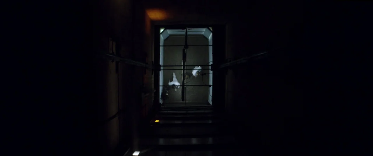 Officer Downe (2016) movie still: establishing — A dark, industrial elevator shaft is framed from a high vertical perspective looking down. Two…