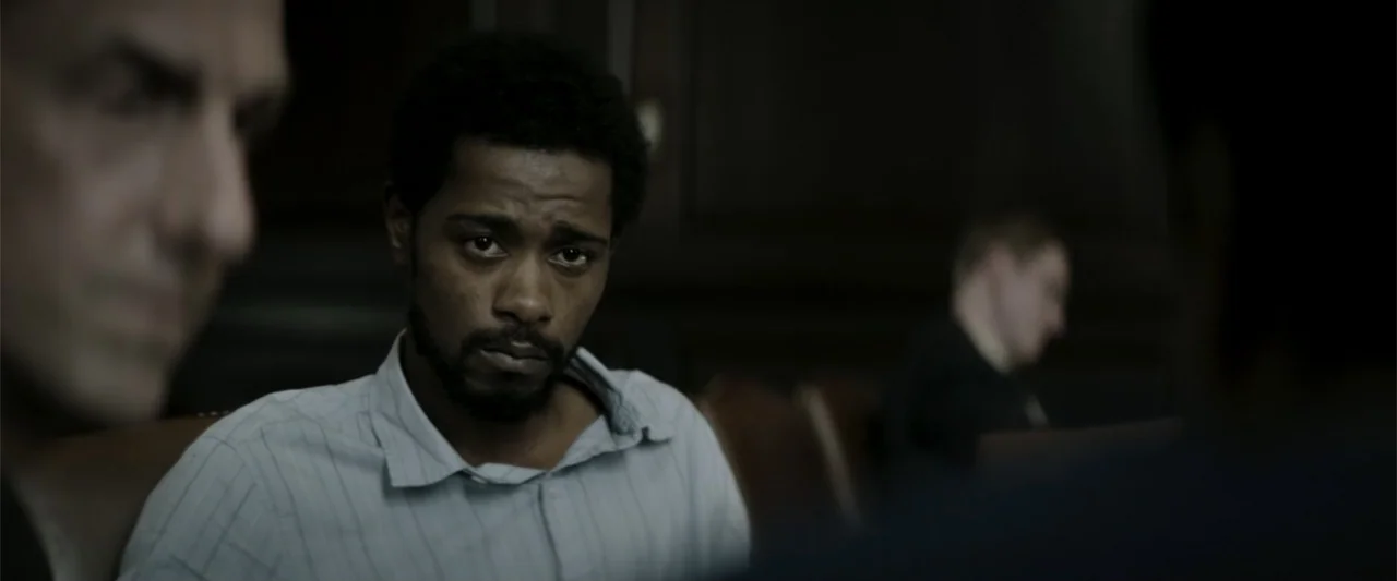 Crown Heights (2017) movie still: medium — A dark, wood-paneled room serves as a setting for a professional meeting. A young man sits at the table…