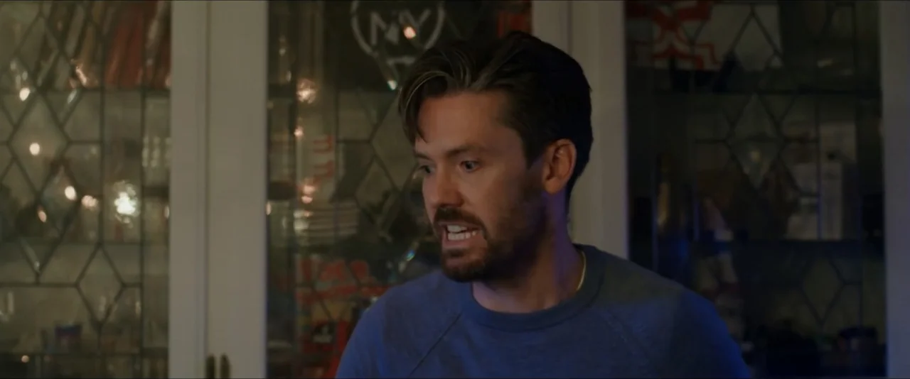 Gatlopp: Hell of a Game (2022) movie still: medium — A dimly lit room features a glass-paned cabinet door in the background. A man in a blue sweater…