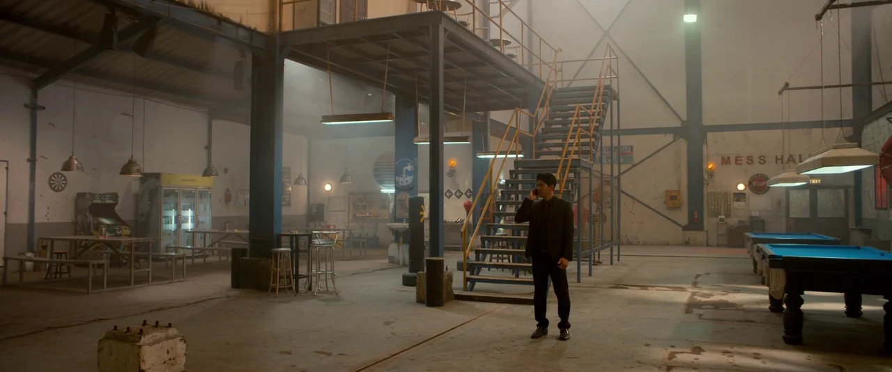 The Night Comes for Us (2018) movie still: fullbody — A large industrial recreation hall features a metal mezzanine structure and pool tables. A man…