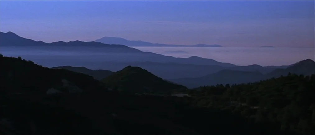 Donnie Darko movie still: establishing — A series of mountain ridges recede into the distance under a clear dawn sky. Low-hanging mist fills the…
