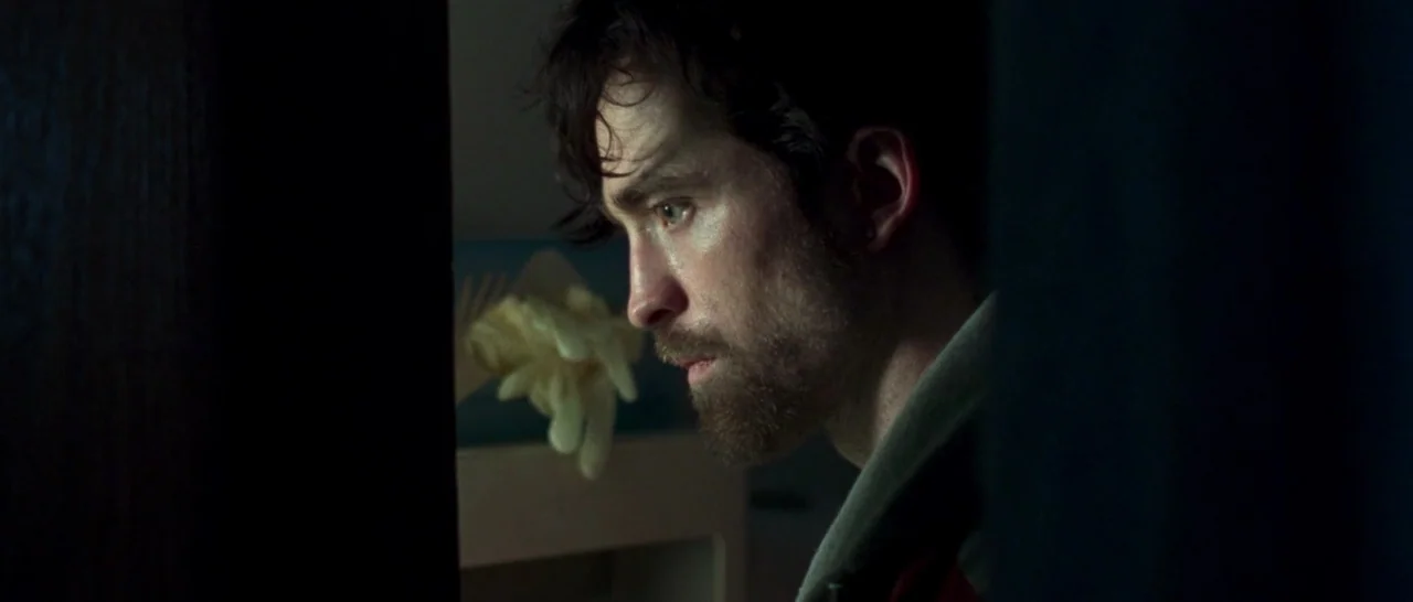 Good Time (2017) movie still: closeup — A man in a dark room peeks through a narrow doorway into a space. He observes something off-camera with a…
