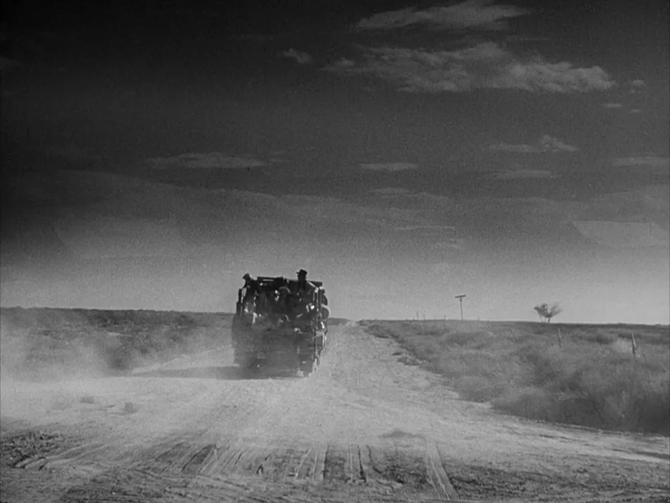 The Grapes of Wrath (1940) movie still: wide — A flat, arid desert landscape spans the horizon under a vast, dark, cloudy sky. A small, open-topped…