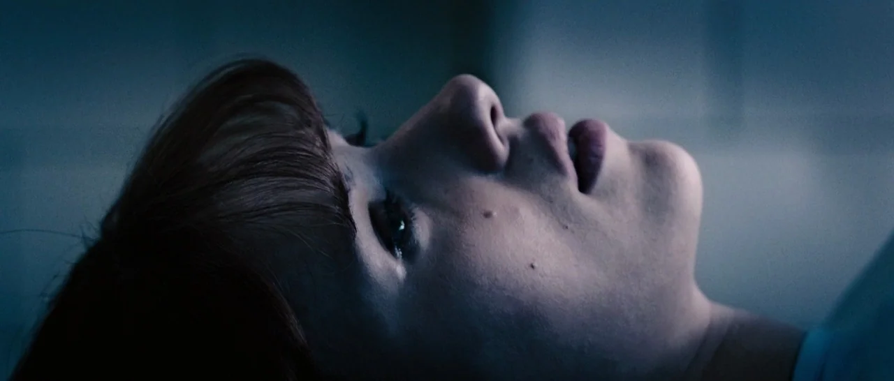 Avengers: Age of Ultron (2015) movie still: closeup — A dimly lit room with blue hues cast across a clinical setting. A woman reclines on a surface…