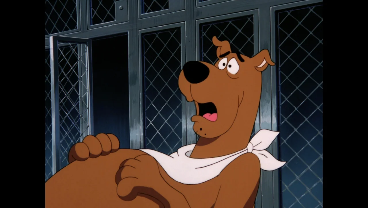 Scooby-Doo on Zombie Island (1998) movie still: medium — A Great Dane sits in front of a metal chain-link enclosure. The dog has a white bandana and…