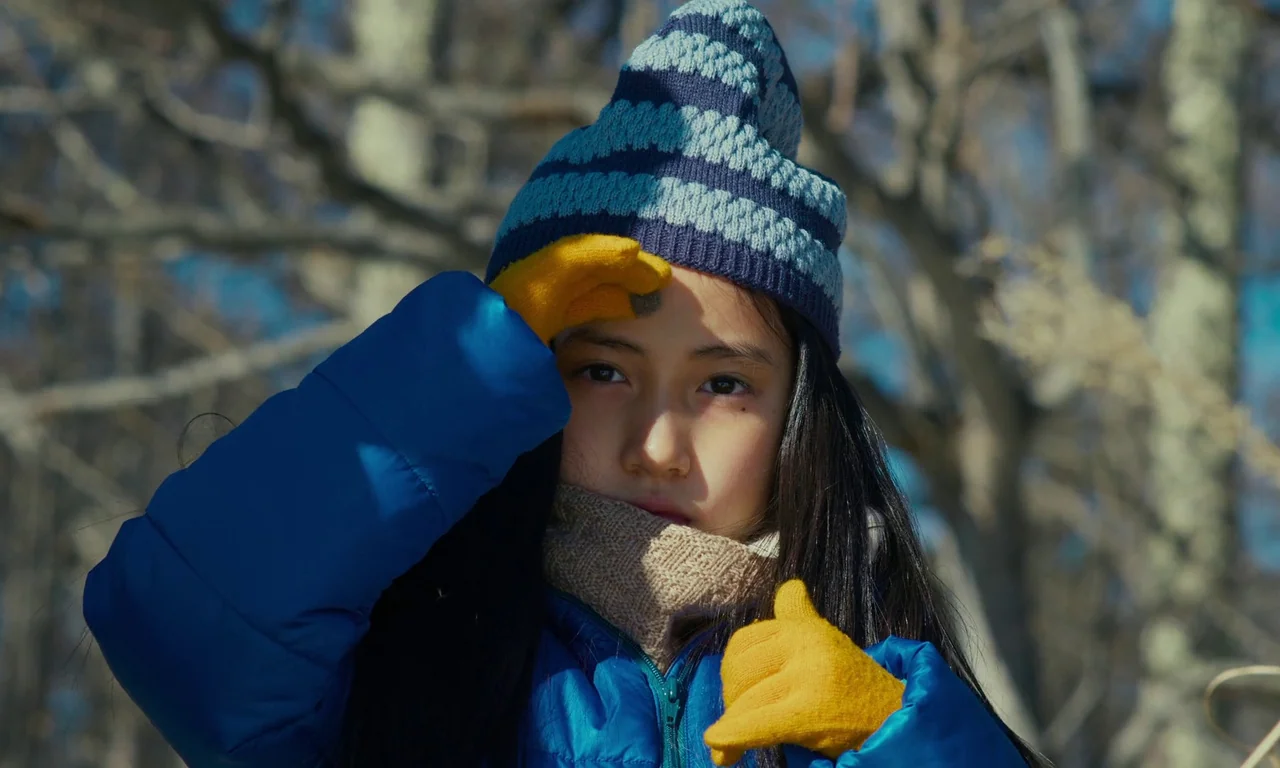 Evil Does Not Exist (2023) movie still: closeup — A girl stands outdoors with leafless, blurred trees visible in the background. She wears a winter…
