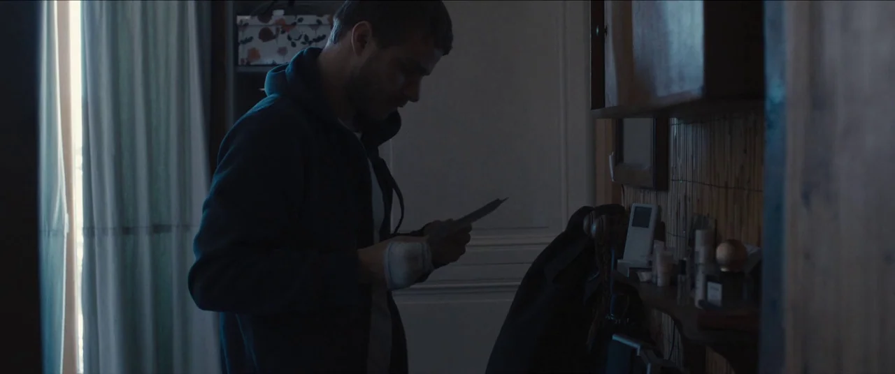 Simon Killer movie still: medium — A dimly lit room with a shelf filled with personal objects and a doorway. A man stands in profile, holding a…