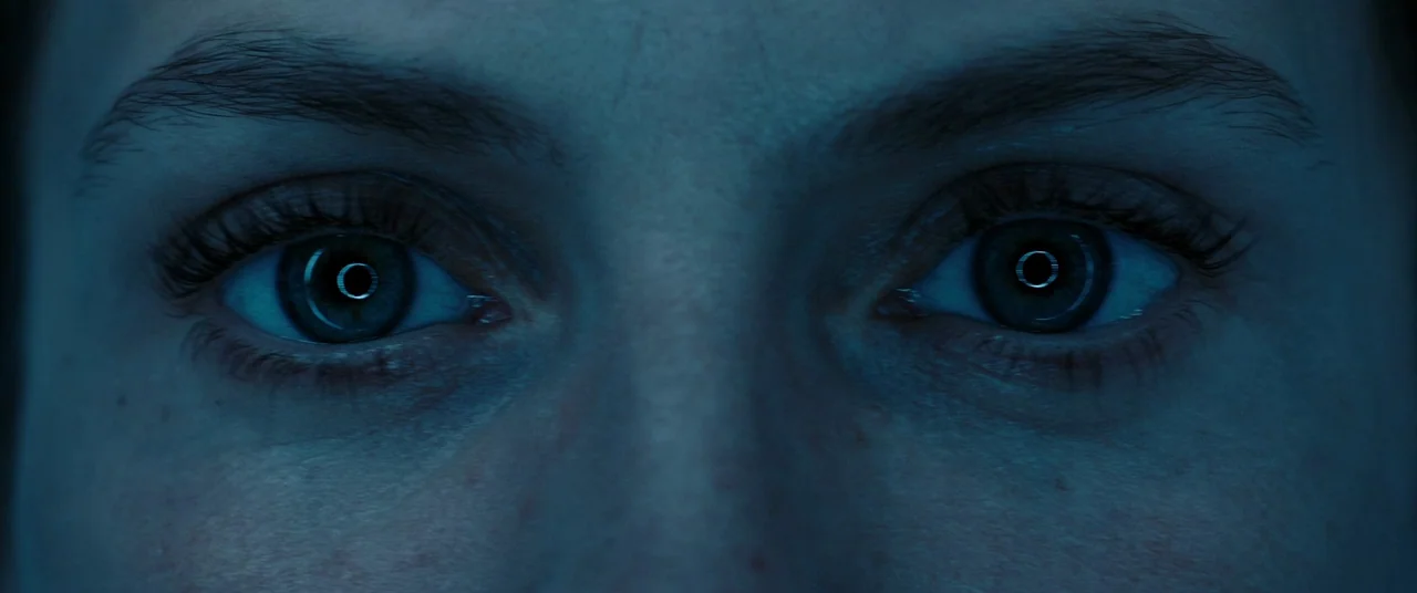 Oxygen (2021) movie still: closeup — A close-up shot focuses on a person's eyes in a dark environment. The eyes reflect a bright ring light or…