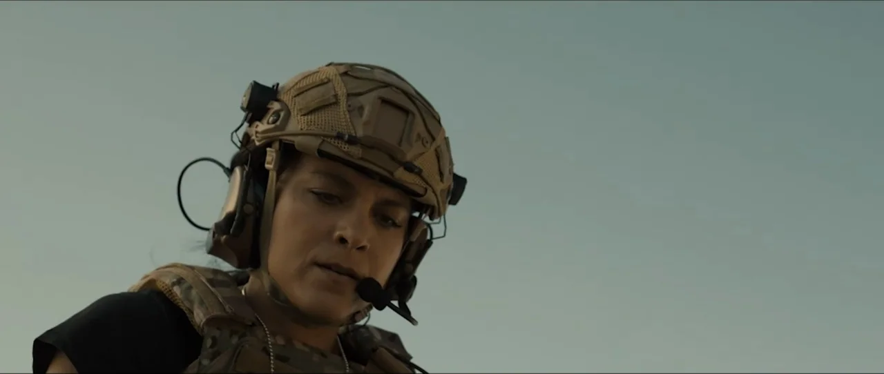 American Sicario (2021) movie still: medium — An open landscape with a clear, pale sky serves as the background. A woman in tactical military gear…