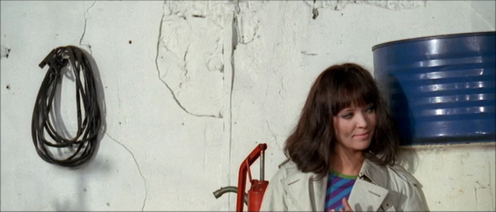 Made in U.S.A. (1966) movie still: medium — A white painted wall with cracks displays a hanging black rubber cable. A woman stands in front of the…