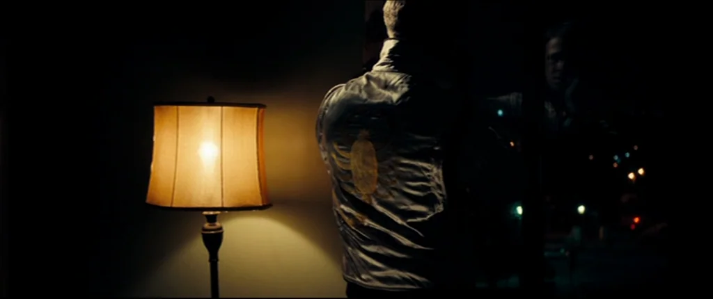 Drive (2011) movie still: medium — A darkened room contains a lit bedside lamp and a man standing before a mirror. The man wears a distinctive white…