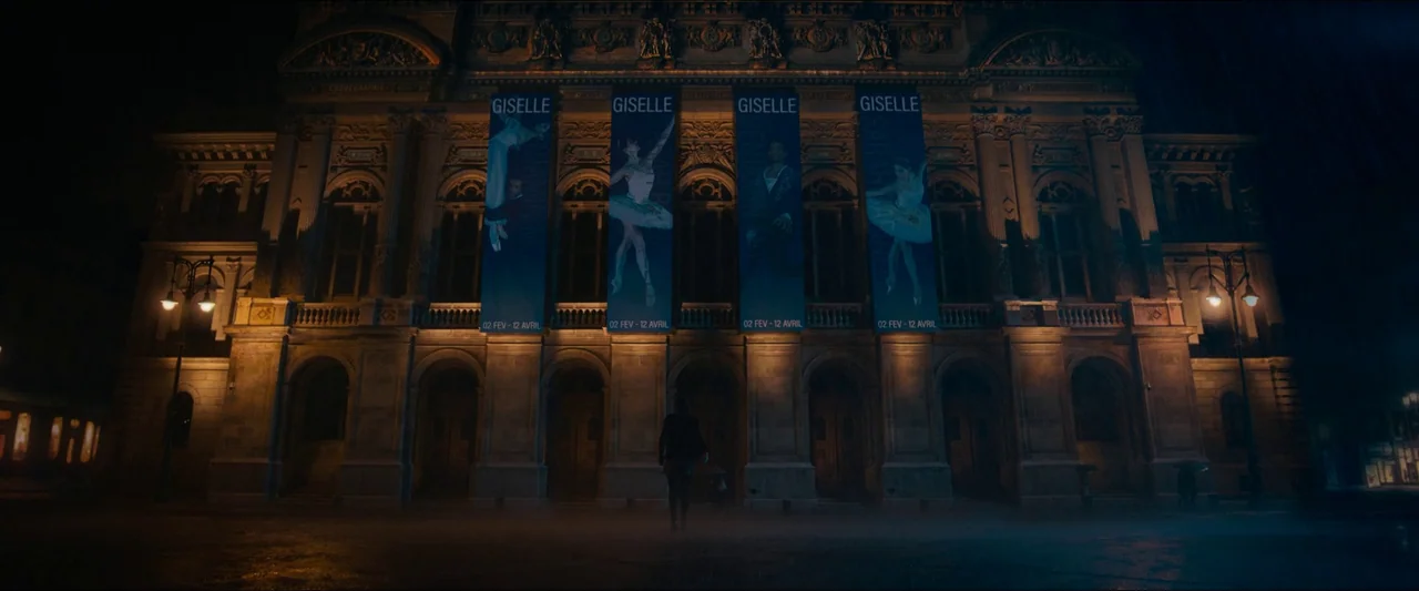 Birds of Paradise (2021) movie still: wide — The night exterior of a grand stone opera house with ornate architectural details. A lone figure walks…