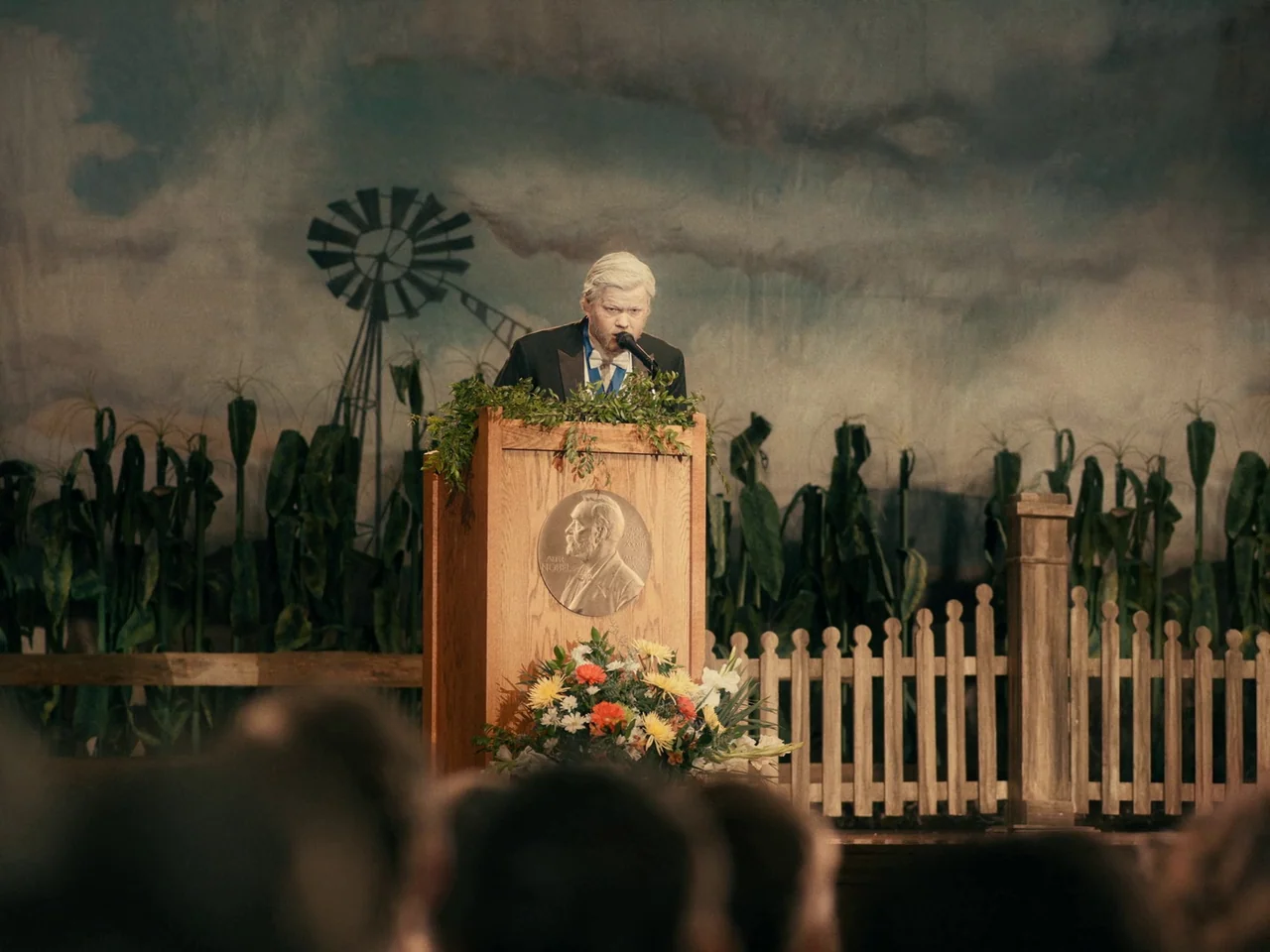 I’m Thinking of Ending Things (2020) movie still: medium — An auditorium stage features a painted backdrop of a farm with a windmill. A man in a…