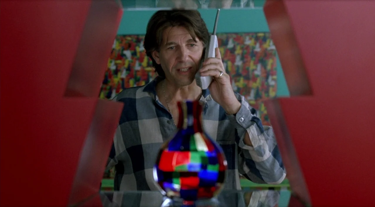 Kika (1993) movie still: medium — A man stands behind a glass shelf framed by bright red geometric partitions. He holds a vintage mobile phone to…