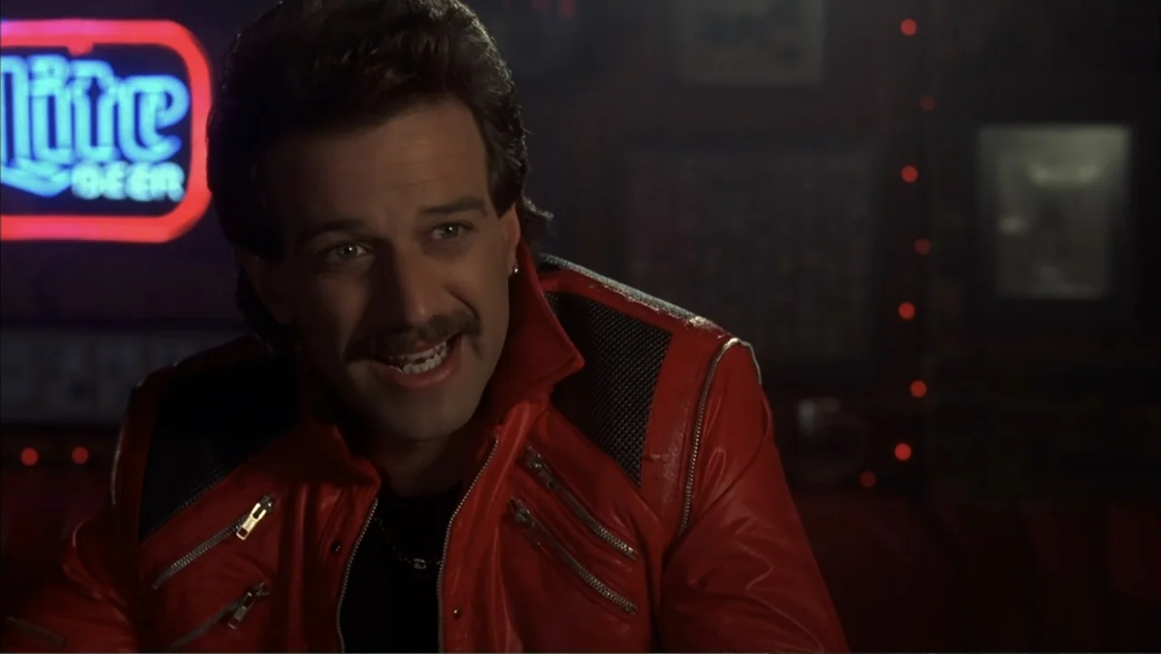 The Wedding Singer (1998) movie still: medium — A dark tavern interior features a glowing neon beer sign mounted on the wall. A man in a red leather…