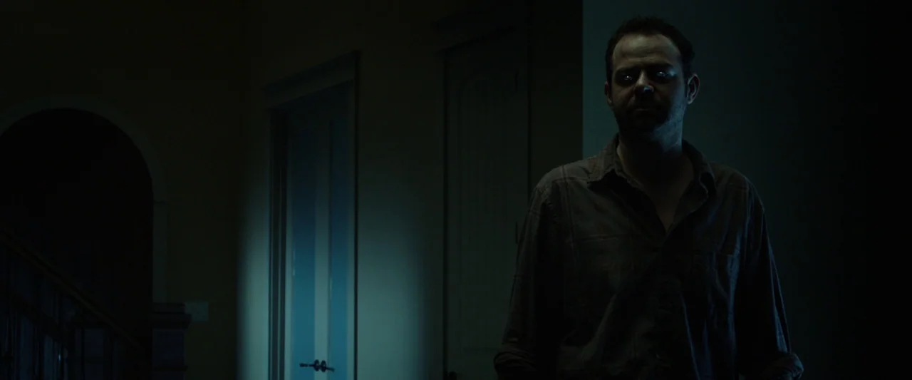 Oculus (2013) movie still: medium — A dimly lit residential hallway at night features closed doors and a dark stairwell. A man stands in the…