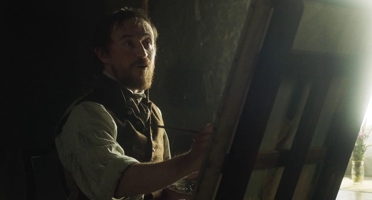 Peterloo (2018) movie still: medium — A dimly lit room contains an artist at work on a wooden easel. The man focuses on applying paint to the canvas…