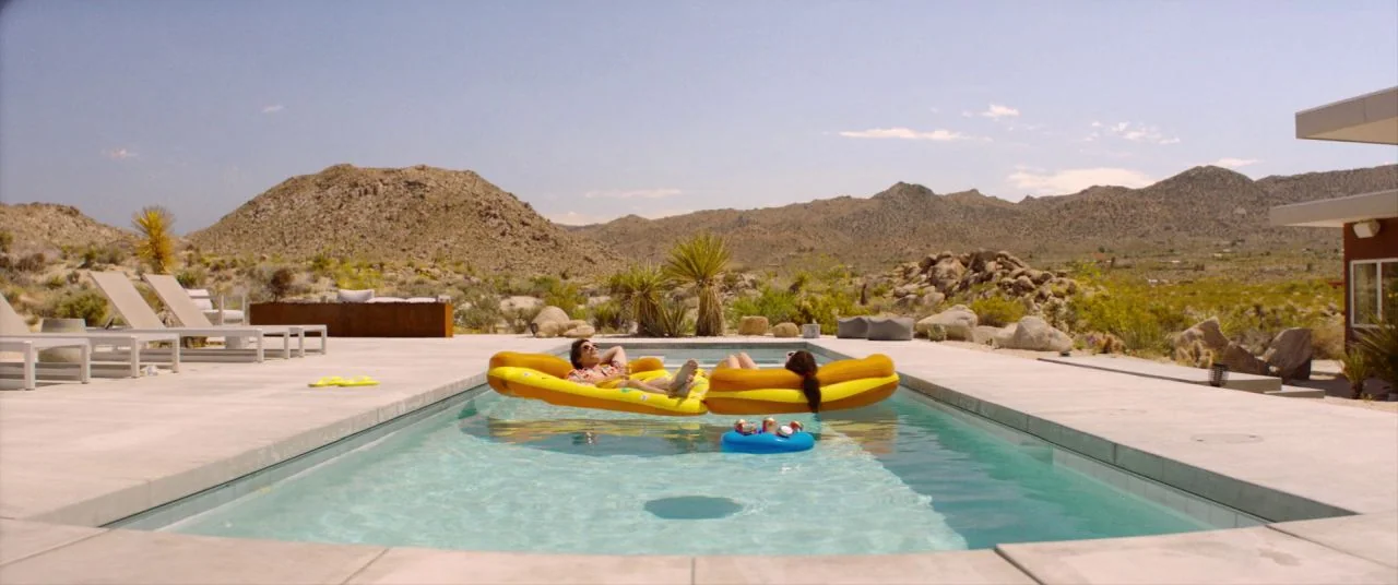 Palm Springs (2020) movie still: wide — A modern pool sits amidst a arid, rocky desert landscape with mountains in the distance. Two people rest on…