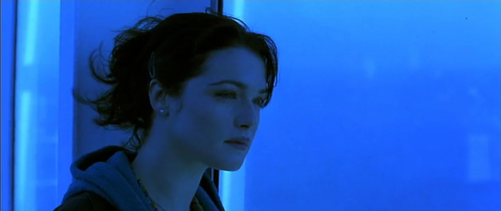 I Want You (1998) movie still: closeup — A person stands near a reflective glass surface illuminated by blue light. The subject gazes ahead while…
