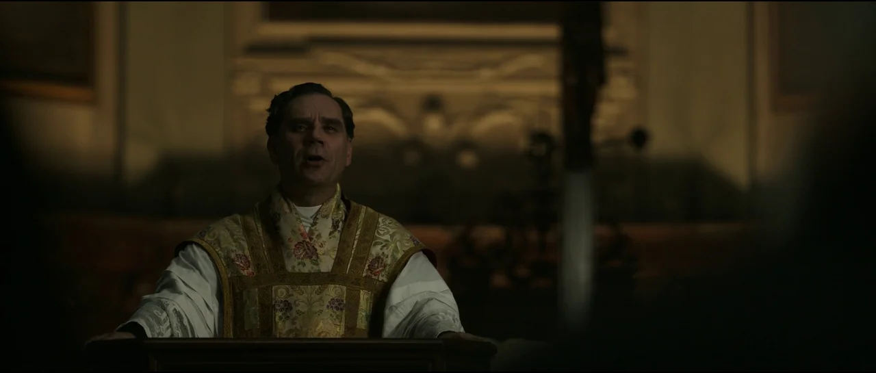 Ferrari (2023) movie still: medium — A dimly lit interior of a stone church features an altar in the background. A priest stands behind a wooden…