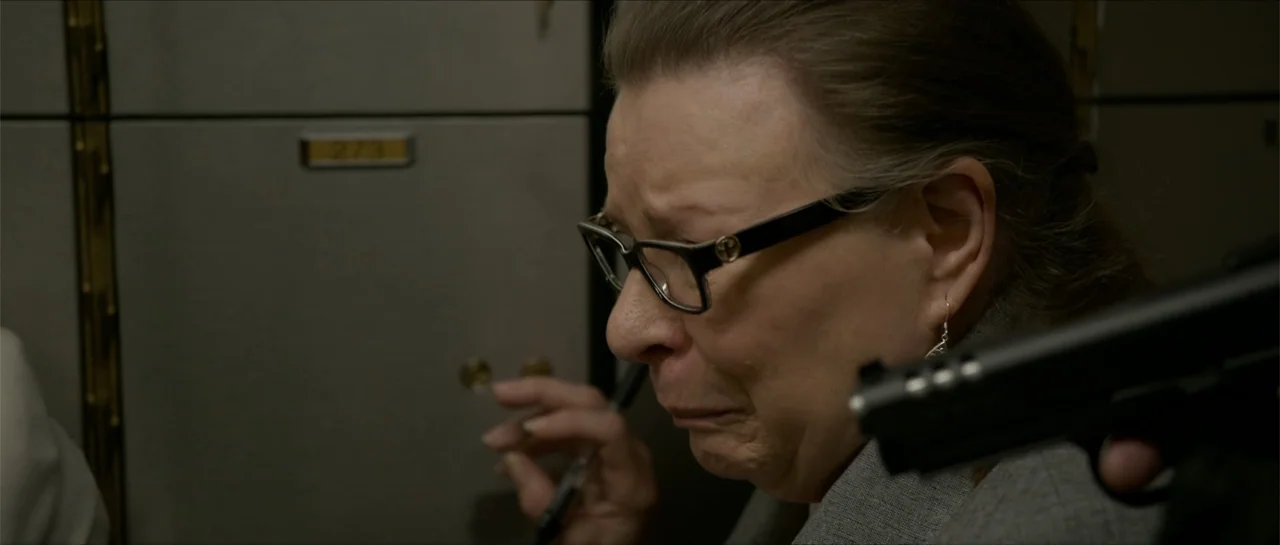 The Vault (2017) movie still: closeup — A dimly lit bank vault contains rows of numbered metal safe deposit boxes. An elderly woman stands near the…