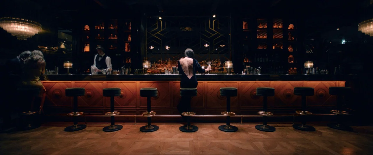 The Substance (2024) movie still: wide — A dimly lit upscale bar features a long wooden counter and rows of stools. A woman in a black backless…