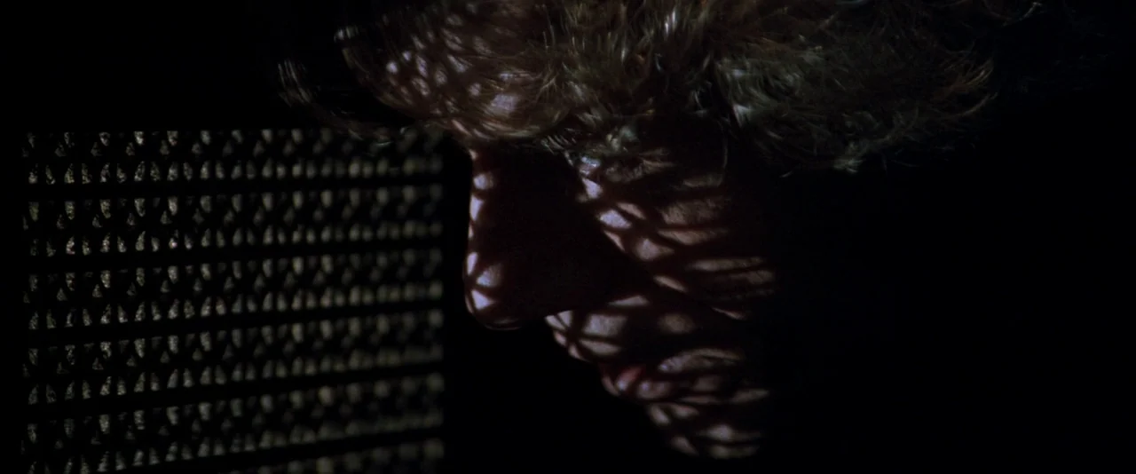 In The Mouth of Madness (1994) movie still: closeup — A dark, dimly lit room contains a person illuminated by narrow beams of light. The subject is…