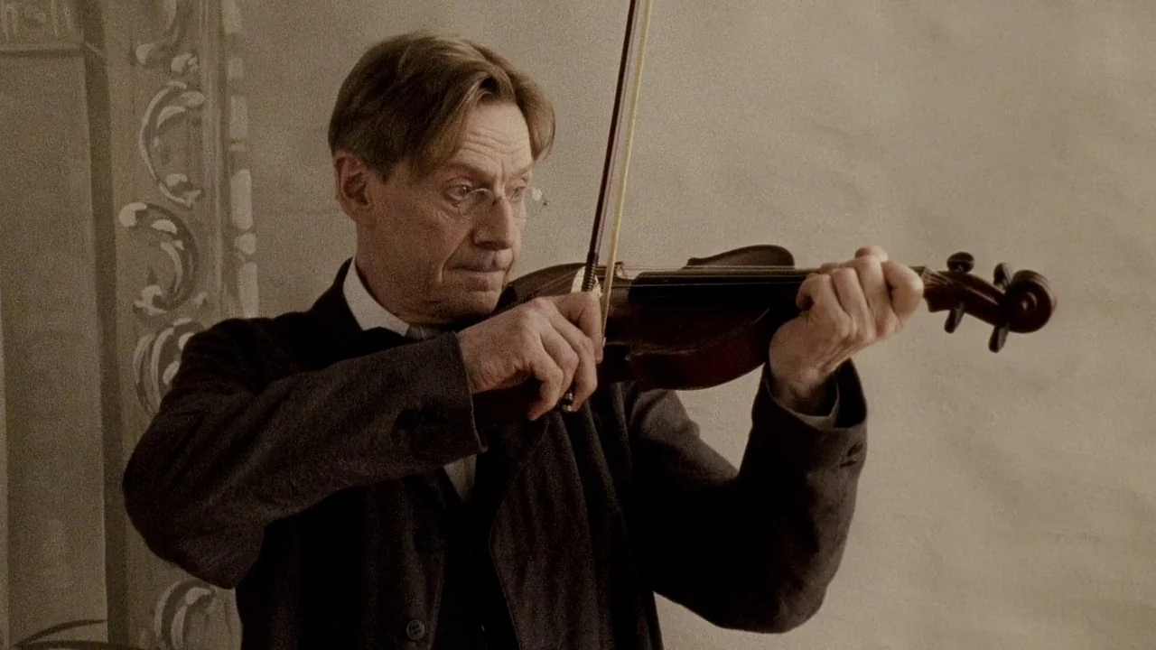 Everlasting Moments (2008) movie still: medium — A man stands in an interior room with a textured light-colored wall. He holds a violin and bow,…
