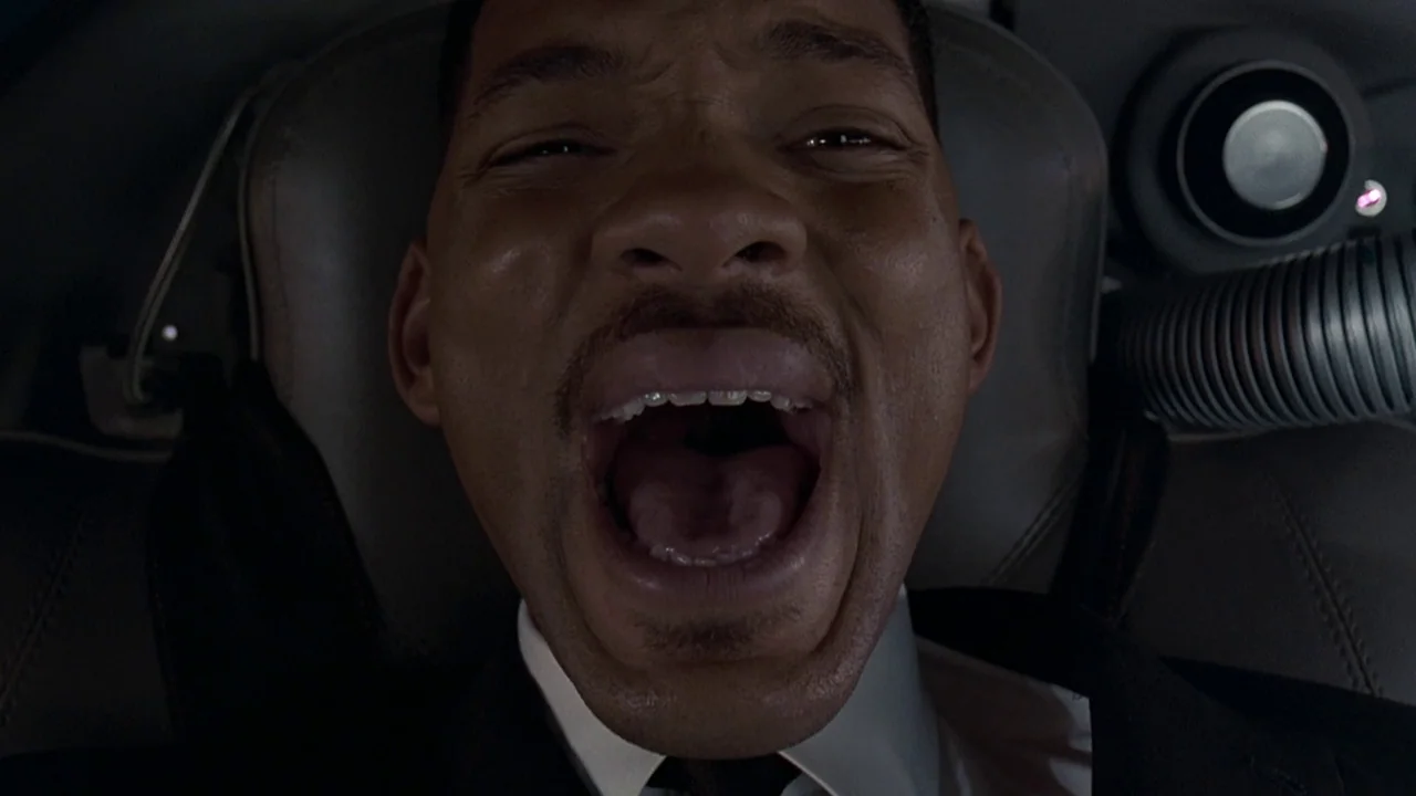 Men In Black (1997) movie still: closeup — A dark vehicle interior with a textured headrest and a coiled metallic cable. A man wearing a suit yells…