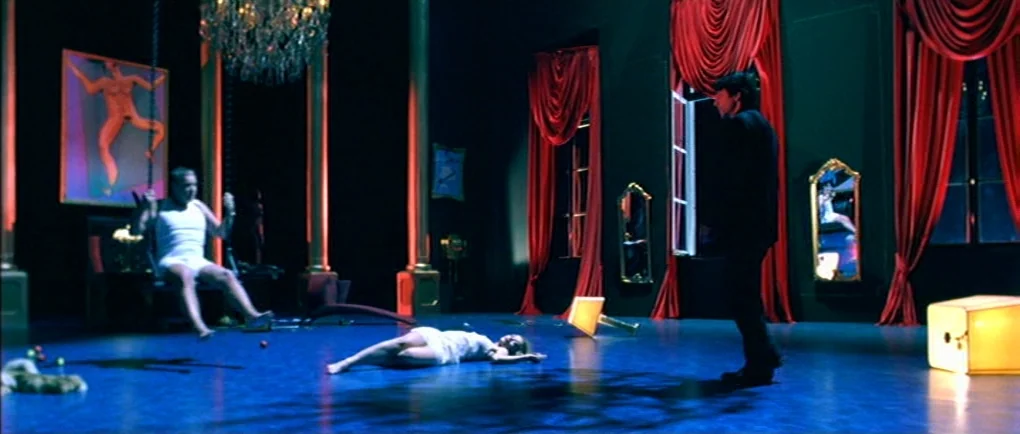 Mortal Transfer (2001) movie still: wide — A theatrical room features dark walls, heavy red curtains, and polished blue floors. Three people occupy…