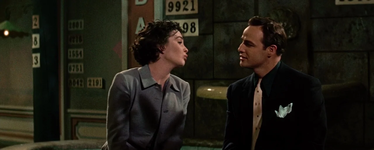 Guys and Dolls (1955) movie still: medium — A dark indoor space features a wall covered with numbered metal placards. A man and a woman sit facing…