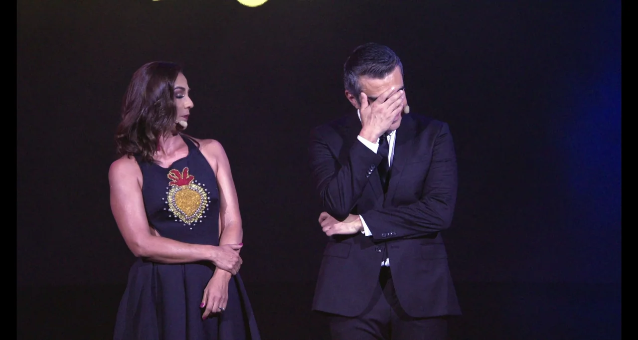 Paired Up (2019) movie still: medium — A dark stage with two presenters standing side by side under bright lights. A woman in a dark dress looks…