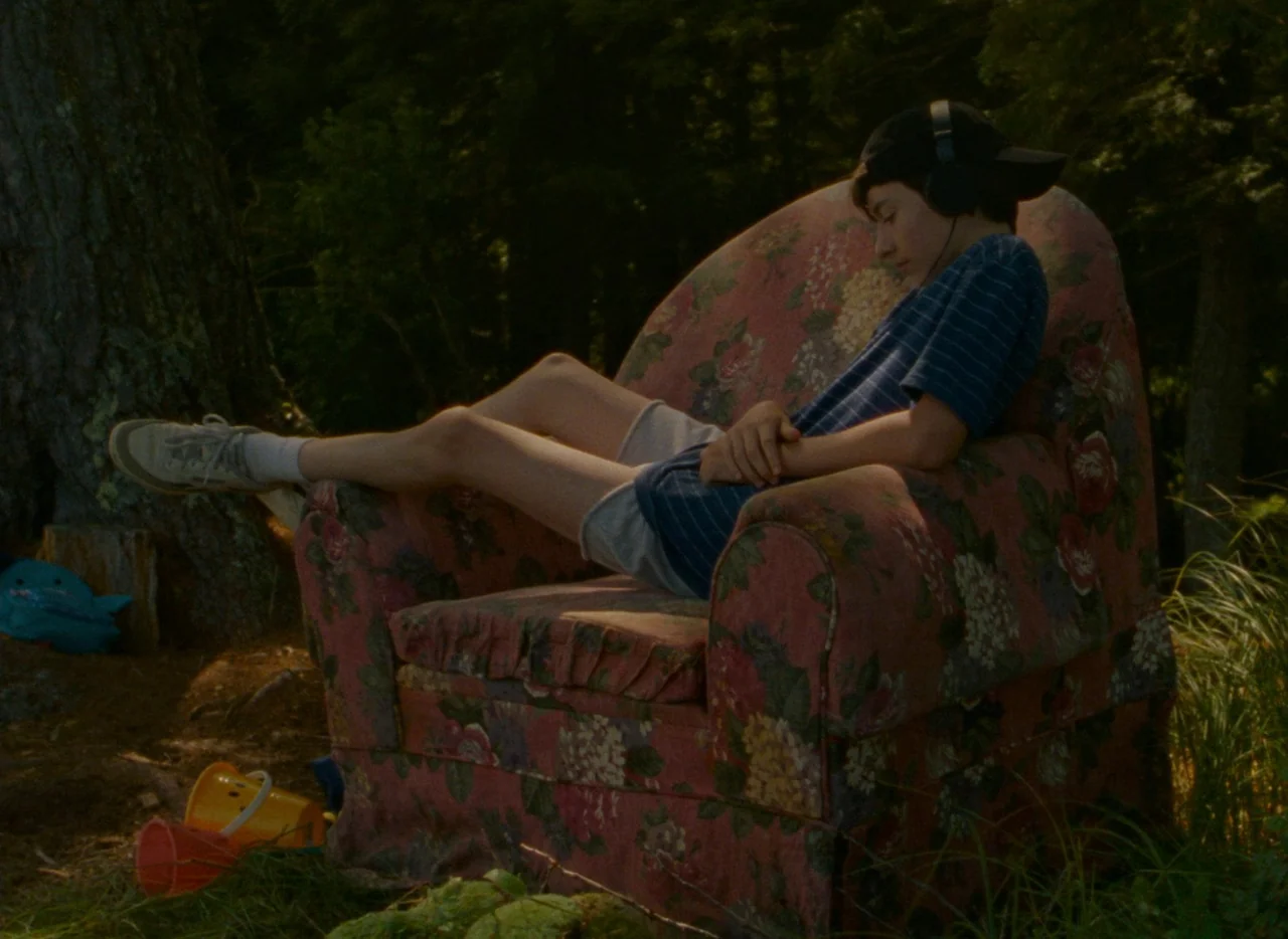 Falcon Lake (2022) movie still: medium — A worn floral-patterned armchair sits outside in a shaded forest clearing. A teenager rests in the chair…