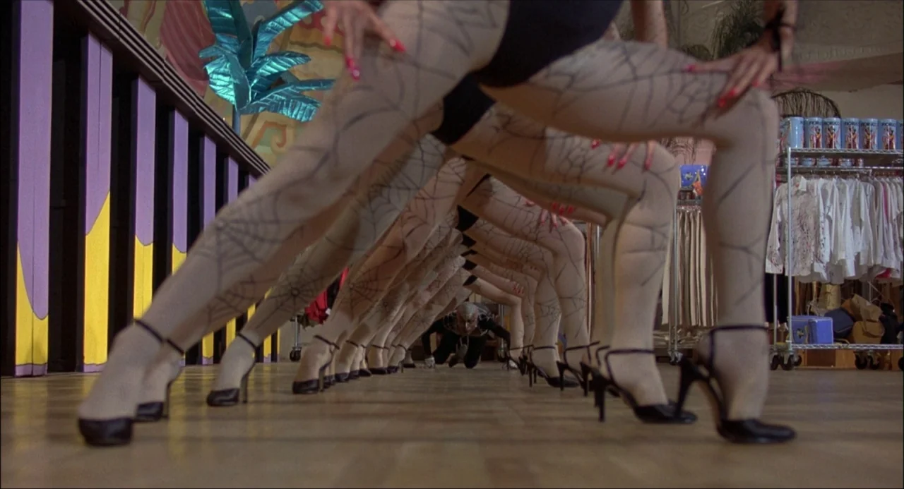 Xanadu (1980) movie still: medium — A line of people in black leotards and patterned hosiery stands in a boutique. The dancers stretch their legs to…