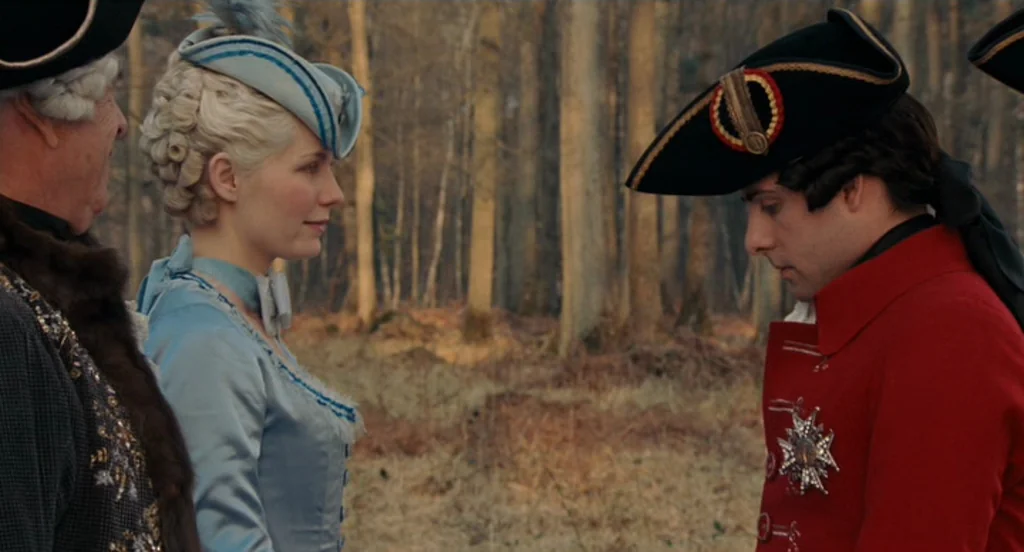 Marie Antoinette (2006) movie still: medium — A wooded area with bare trees forms the background for two figures. A woman in blue and a man in a red…