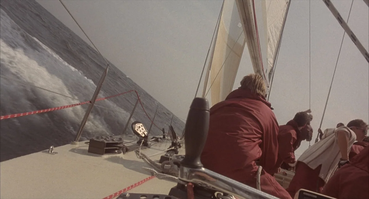 Masquerade (2023) movie still: wide — A sailboat deck is heavily tilted on a choppy sea under a pale sky. Several crew members in matching red…