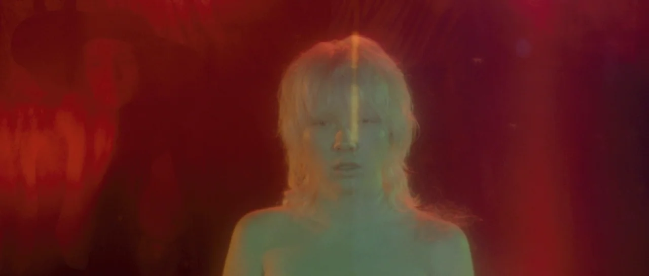After Blue (2021) movie still: medium — A person with blonde hair stands in a dimly lit, nondescript dark environment. A bright vertical beam of…