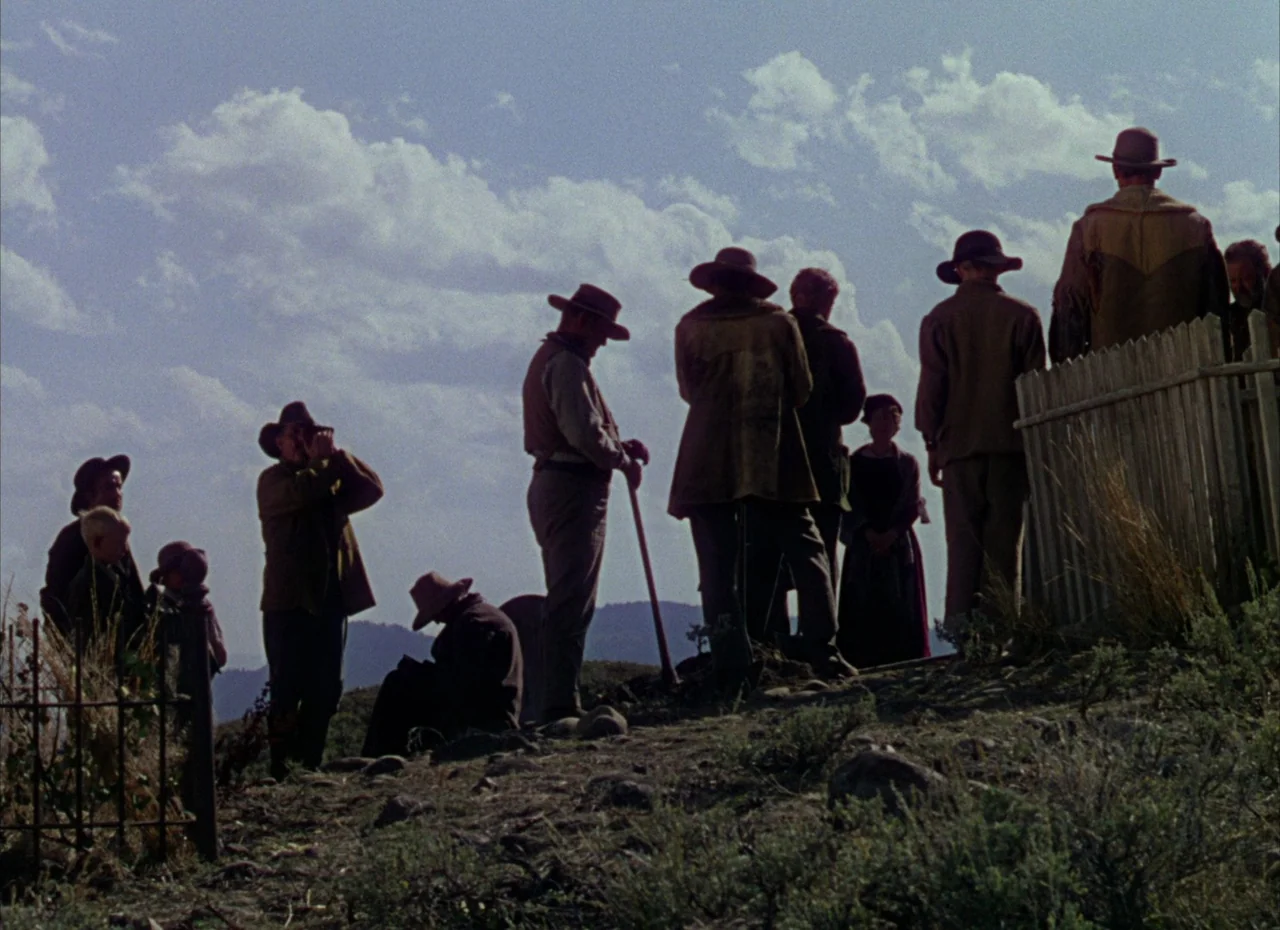 Shane (1953) movie still: fullbody — A group of people wearing 19th-century western attire gather at a hillside cemetery. They stand silently in a…