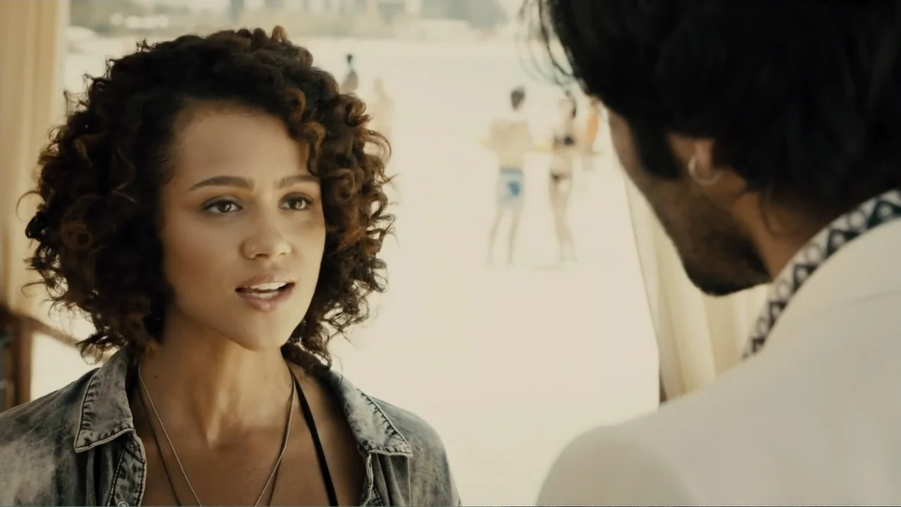 Furious 7 (2015) movie still: medium — A sunny beach cabana setting with a soft focus background of the shore. A woman in a denim shirt engages in…