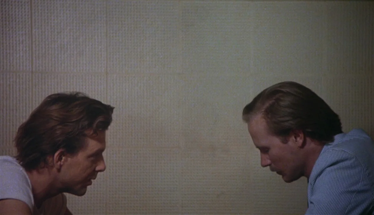 Body Heat (1981) movie still: medium — Two men sit facing each other against a plain, textured wall in a room. The subjects engage in a conversation…