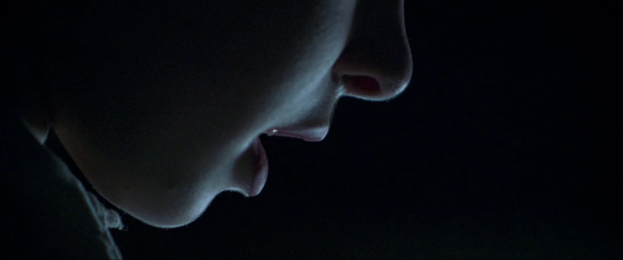 The Wonder (2022) movie still: closeup — A close-up profile view of a face set against a pitch-black background. Only the edge of the nose, lips,…