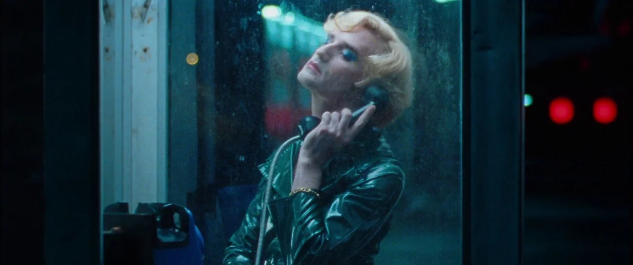 Knife+Heart (2018) movie still: medium — A person stands inside a glass-walled booth at night with city lights behind them. The individual wears a…