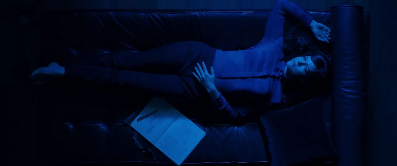 Elizabeth Harvest movie still: fullbody — A woman lies on a leather sofa in a dark room under blue light. An open notebook with handwritten text…