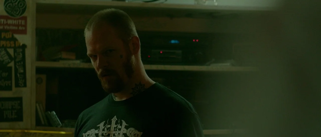 Green Room (2015) movie still: medium — A dimly lit room with walls covered in stickers and a shelf containing electronic equipment. A man with a…