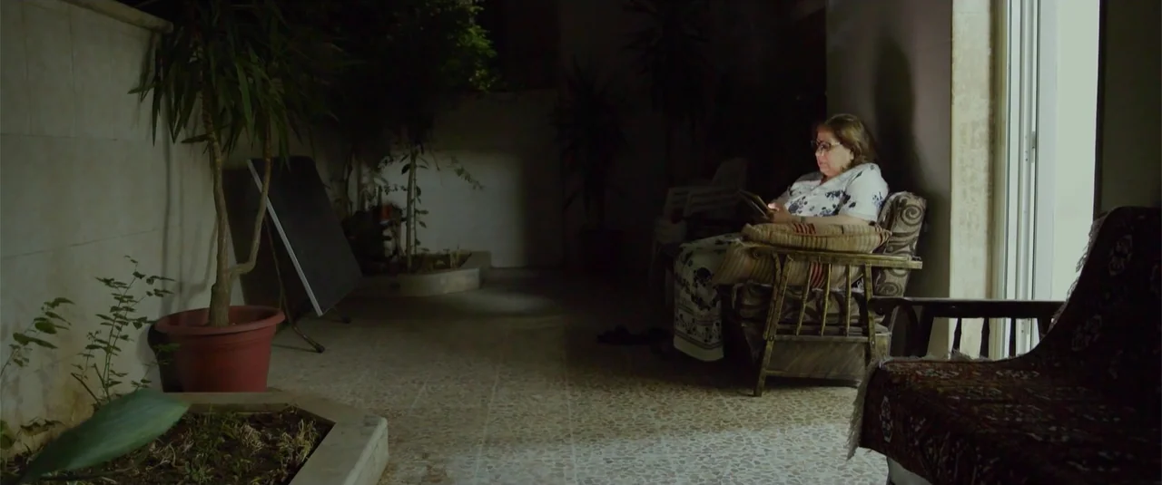 Simple as Water (2021) movie still: medium — A dimly lit outdoor stone patio area features potted plants and various chairs. An older woman sits in…