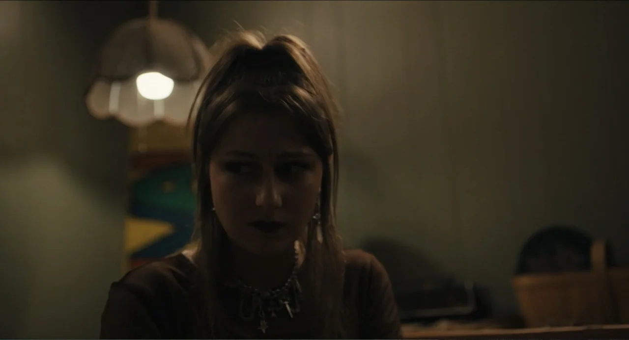 Hell of a Summer (2025) movie still: closeup — An interior space is dimly lit by a singular overhead decorative pendant lamp. A young woman with a…