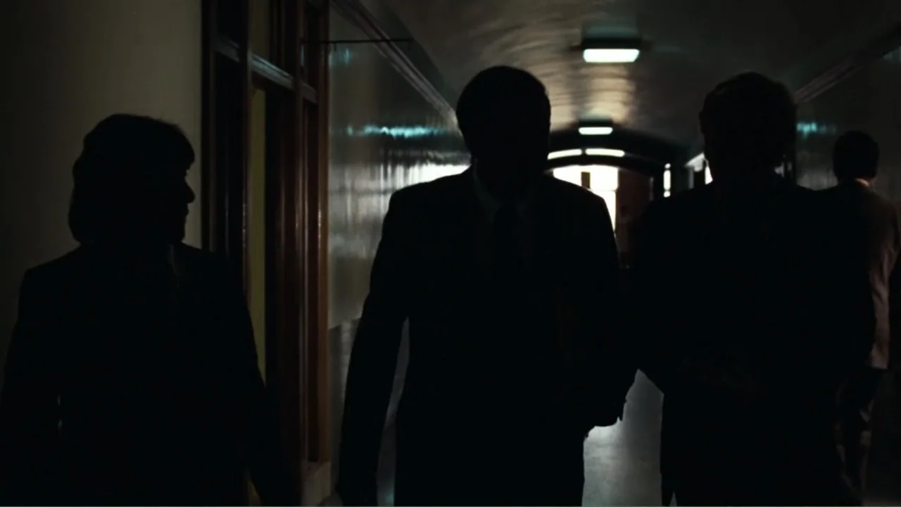 All the President's Men (1976) movie still: medium — A long, dimly lit institutional hallway has figures walking in silhouette toward the camera.…